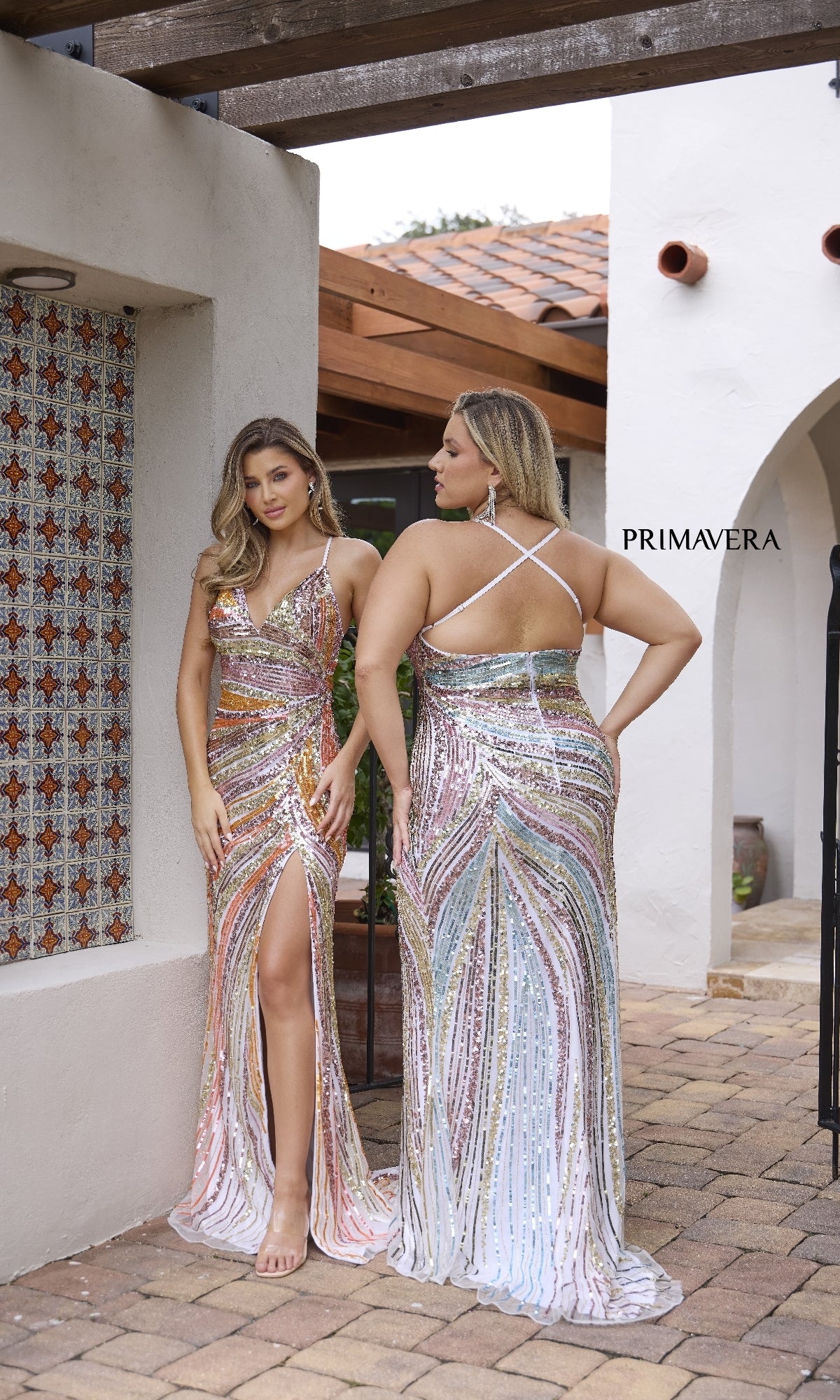 Two women pose outdoors in identical Primavera 4302 Strappy-Back Sequin Prom Gowns featuring deep v-necks, side slits, and crisscross straps—one faces forward as the other shows off the gown’s striking back design.